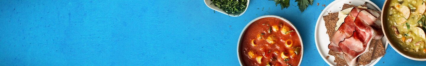 Diverse gerechten op een blauwe achtergrond, inclusief soep, brood met vleeswaren en groente.