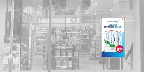 Reclameposter voor Sensodyne tandpasta voor gevoelige tanden, met een aanbieding voor twee tubes voor 8 euro, zichtbaar in een etalage van een winkel.