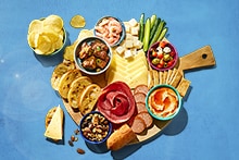 Borrelplank met verschillende hapjes zoals chips, garnalen, vleeswaren, kaas, groentesticks, crackers, bruschetta, olijven, dips en noten.