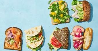 Verschillende soorten gezonde belegde broodjes en sandwiches, waaronder volkorenbrood met zalm en kiemen, flatbread met groenten, brood met kaas en tomaat, sandwich met runderlappen en ingelegde ui, en broodjes met avocado en sla, gepresenteerd op een blauwe ondergrond.