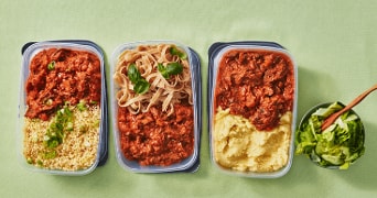 Drie plastic bakjes met verschillende maaltijden, waaronder couscous met tomatensaus en lente-ui, tagliatelle met tomatensaus en basilicum, en aardappelpuree met tomatensaus. Daarnaast een bakje met een groene salade en houten lepel.