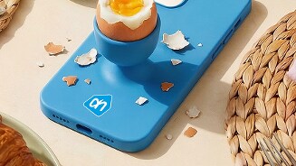 Een gekookt ei in een eierdop op een felblauwe telefoonhoes met Albert Heijn-logo, omringd door gekleurde paaseieren en gevlochten placemats.