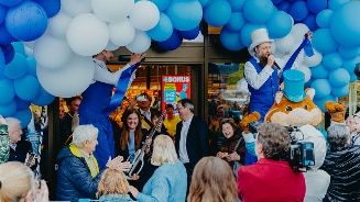 Opening van een Albert Heijn winkel met blauwe en witte ballonnen, entertainers in blauwe kostuums met hoge hoeden en de bekende hamster mascotte, omringd door een groep feestvierende mensen.