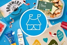 Producten van Albert Heijn, waaronder Brouwers Premium Bier, Caesar kipsalade, watermeloenstukjes, en Dove Invisible Dry deodorant, naast een Red Bull blikje en zonnebrandcrème, rond een canvas tas met feestelijke confetti en zonnebril.