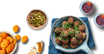 Gehaktballetjes met peterselie geserveerd in een schaal, omringd door gedroogde abrikozen, pistachenoten en twee glazen thee.