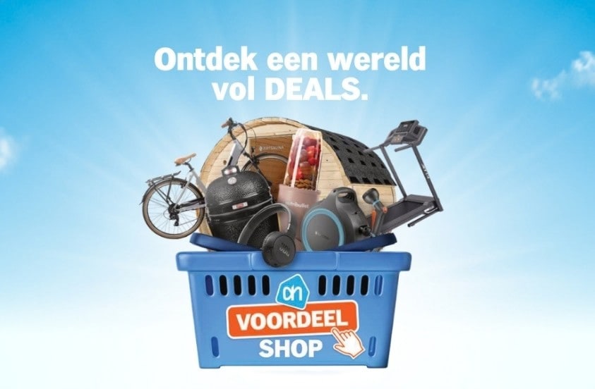 Blauwe Albert Heijn winkelmand met diverse producten zoals een fiets, barbecue, sauna, JBL speaker, NutriBullet blender, loopband en boodschappen, tegen een blauwe lucht met de tekst "Ontdek een wereld vol DEALS" en het logo van AH Voordeel Shop.