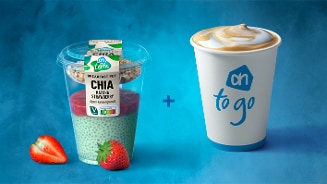 Ontbijtpot met AH Chia matcha aardbei en granola naast een beker koffie van AH to go, op blauwe achtergrond met aardbeiën op de voorgrond.