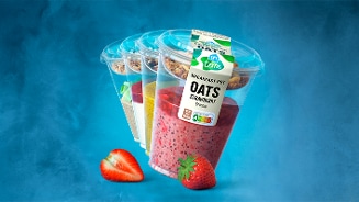 Ontbijtpotten van AH Terra met granola en verschillende smaken, waaronder aardbei, gepresenteerd op een blauwe achtergrond met verse aardbeien op de voorgrond.