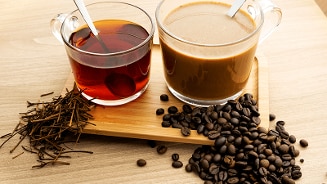 Glazen kop thee en kop koffie op een houten dienblad met theebladeren en koffiebonen ernaast.