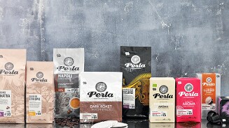 Verschillende verpakkingen van Perla koffie op een rij tegen een grijze achtergrond, inclusief Dark Roast koffiepads en diverse soorten bonen.