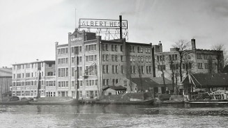 Zwart-witfoto van een historisch Albert Heijn gebouw met een grote reclamebord en boten op een kanaal.