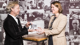 Twee personen reiken elkaar een mini winkelwagentje aan voor een muur met historische foto's van Albert Heijn.