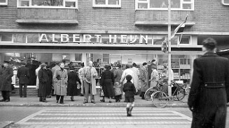 Zwart-wit foto van mensen die zich verzamelen voor een Albert Heijn winkel in de jaren '50.