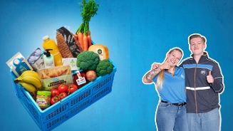Albert Heijn winkelmand gevuld met verse groenten, fruit, brood en zuivelproducten, naast twee medewerkers in Albert Heijn kleding tegen een blauwe achtergrond