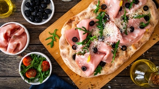 Focaccia belegd met mortadella, olijven, rucola en Parmezaanse kaas op een houten plank, met schalen zwarte olijven, rucola en cherrytomaten erbij.