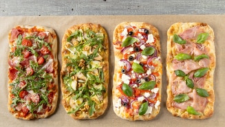 Vier verschillende soorten rechthoekige pizza's met diverse toppings op een houten ondergrond: tomaat en rucola, artisjok en walnoot, olijf en basilicum, ham en basilicum.