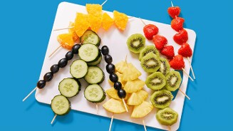 Verschillende spiesjes met vers fruit en komkommer op een witte snijplank, waaronder aardbeien, kiwi, ananas, sinaasappel, druiven en komkommer, op een blauwe ondergrond.