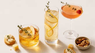 Drie verschillende cocktails, elk gegarneerd met kruiden en fruit, geserveerd naast snacks zoals amandel, walnoot, stukjes kaas en toast met roomkaas.