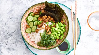 Pokébowl met witte rijst, komkommer, edamame, zeewiersalade, krokante stukjes, nori en sojasaus, geserveerd met eetstokjes.