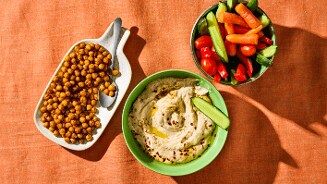 Schaaltje geroosterde kikkererwten, een kom hummus met olijfolie en kruiden, en een bakje met verse groente zoals wortels, paprika en komkommer, op een oranje tafelkleed.
