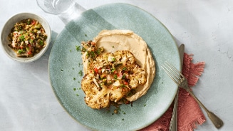 Geroosterde bloemkoolsteak met hummus en salsa op een groen bord, geserveerd met bestek en een klein bakje extra salsa.