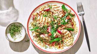 Quinoasalade met gegrilde halloumi, amandelen, radijs, sugarsnaps en tuinkers, geserveerd op een wit bord met rode rand.