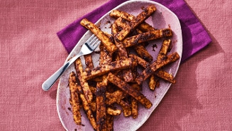 Krokante tempeh frietjes op een ovaal bord, geserveerd met een vork op een roze en paarse ondergrond.