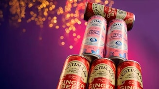 Een stapel Fentimans Ginger Beer en Rose Lemonade blikjes tegen een paarse achtergrond met lichtbollen.