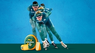 Schaatsers in groene pakken racen voor een blauwe achtergrond, naast verschillende stukken en een wiel Zaanlander kaas van Albert Heijn.