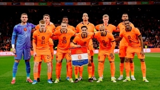 Voetbalteam in oranje tenue poseert op het veld voor een wedstrijd, met een vlag van de Koninklijke Nederlandse Voetbalbond (KNVB).