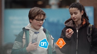 Twee tieners buiten; één houdt een flesje water vast met een Albert Heijn-logo, de ander eet een appel met een KNVB-logo zichtbaar.