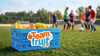 Blauwe krat met de tekst "Team fruit" van Albert Heijn gevuld met bananen, appels, sinaasappels en druiven op een voetbalveld, met op de achtergrond voetballende kinderen.
