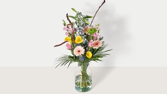 Gemengde boeket bloemen in een glazen vaas, waaronder gele calla lelies, roze gerbera's en groene bladeren.