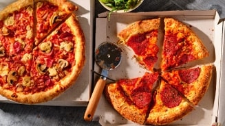 Twee verschillende pizza's in geopende dozen, met een pizzasnijder tussenin. Links een pizza met champignons en ham, rechts een pizza met pepperoni.