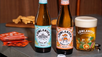 Lowlander 0.0% Wit en Mango I.P.A. bierflessen naast een glas bier en een stapel oranje spelkaarten op een tafel, met toast op de achtergrond