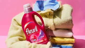 Fleuril Color wasmiddel, fles geplaatst op een stapel kleurrijke kleding tegen een roze achtergrond.