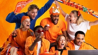 Groep mensen gekleed in oranje kleding, juichend en feestend voor een sportevenement, met oranje accessoires en decoraties op de achtergrond.