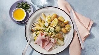 Witte asperges met gekookte krieltjes, plakken ham en peterselie, geserveerd op een bord met bestek, olie en extra kruiden ernaast.