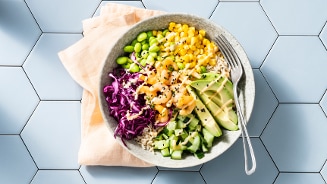 Pokébowl met garnalen, avocado, maïs, rode kool, komkommer, edamame en rijst op een grijze ondergrond.