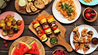 Gevarieerde zomerse barbecuegerechten op een houten tafel, inclusief groenten- en vleesgerechten, meloen en bessen.