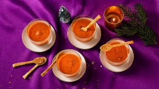 Vier glazen met tomatensoep en een soepstengel, op een paarse ondergrond met een kaars en kersttakje.