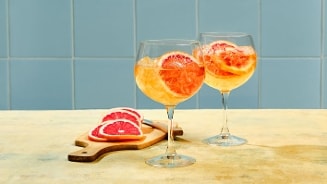 Twee glazen cocktails met schijfjes grapefruit en ijsblokjes, naast een snijplank met verse sinaasappelpartjes.