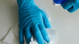 Hand met blauwe handschoen die een oppervlak schoonmaakt met een doek en een spuitfles.