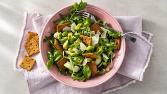 Groene salade met rucola, edamame, crackers en Parmezaanse kaas in een roze kom, geserveerd op een lichtroze servet.