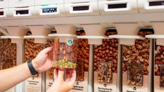 Zelfbedieningsdispenser voor noten en zaden bij Albert Heijn, waarbij een klant pompoenpitten in een potje tapt.