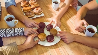 Vier mensen pakken muffins van een bord op een houten tafel met koffie en donuts