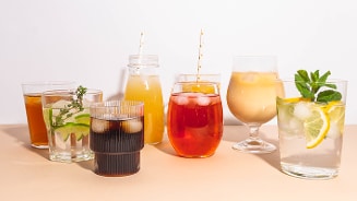 Assortiment van kleurrijke dranken in verschillende glazen, waaronder frisdrank, sap en water met citroen en munt, op een lichte achtergrond