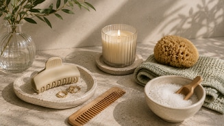 Alt tekst: Luxe spa accessoires op een tafel, waaronder een glazen vaas met takken, een brandende geurkaars in een ribbelglas, een handdoek met een natuurlijke spons, een schaaltje met badzout en houten schepje, een haarclip, sieraden en een houten kam.