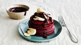 Rode pannenkoeken met plakjes banaan en gesmolten chocolade, geserveerd op een blauw bord met een vork ernaast.