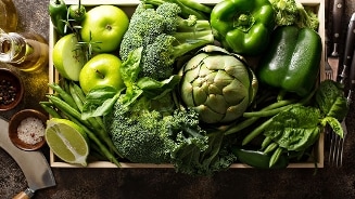 Groene groenten en fruit in een houten kist, waaronder broccoli, groene paprika's, groene appels, artisjok, sperziebonen, basilicum en limoen, met schaal zeezout, pepermolen en olijfolie aan de zijkant.